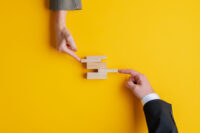 Business teamwork and cooperation concept - hands of businessman and businesswoman pushing wooden peg in a stack of them. Over yellow background, with copy space.